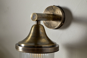 The nkuku Magola Outdoor Wall Lamp in antique brass features a round base, cylindrical arm, flared top, and ribbed glass shade, casting subtle shadows for stylish exterior lighting.
