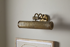 The Tinje Picture Wall Light by nkuku, in antique brass with horizontal ridges and an arched bracket, is mounted above a framed, textured off-white canvas, adding elegant vintage lighting to artwork.