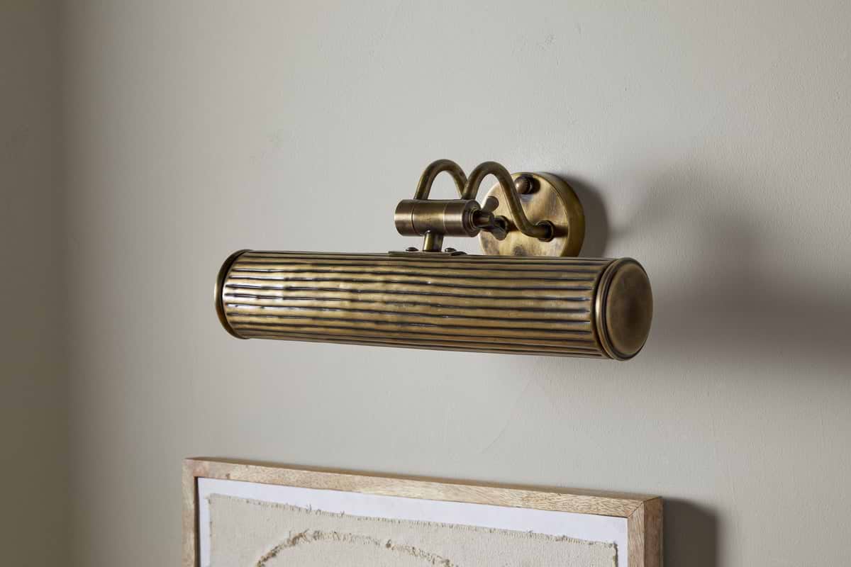 The Tinje Picture Wall Light by nkuku, in antique brass with horizontal ridges and an arched bracket, is mounted above a framed, textured off-white canvas, adding elegant vintage lighting to artwork.