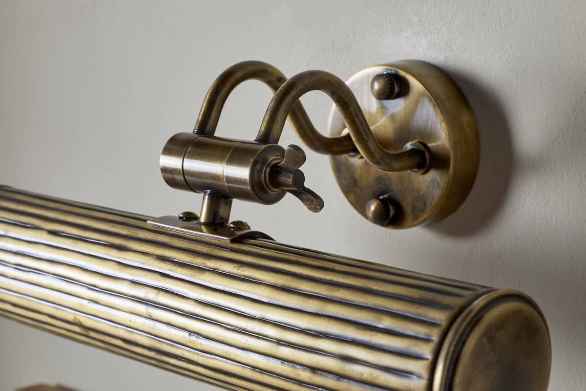 Close-up of nkuku’s Tinje Picture Wall Light in antique brass, featuring a ribbed cylindrical shade, ornate curved arm, and round backplate on a pale wall—perfect for adding elegant lighting to your artwork.