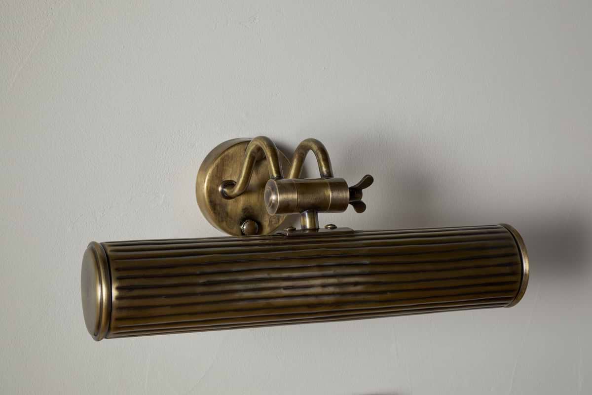 The Tinje Picture Wall Light - Antique Brass by nkuku features a horizontally ribbed cylindrical shade, adjustable arm, and round backplate. Its aged brass finish adds vintage charm, perfect for showcasing artwork on light gray walls.