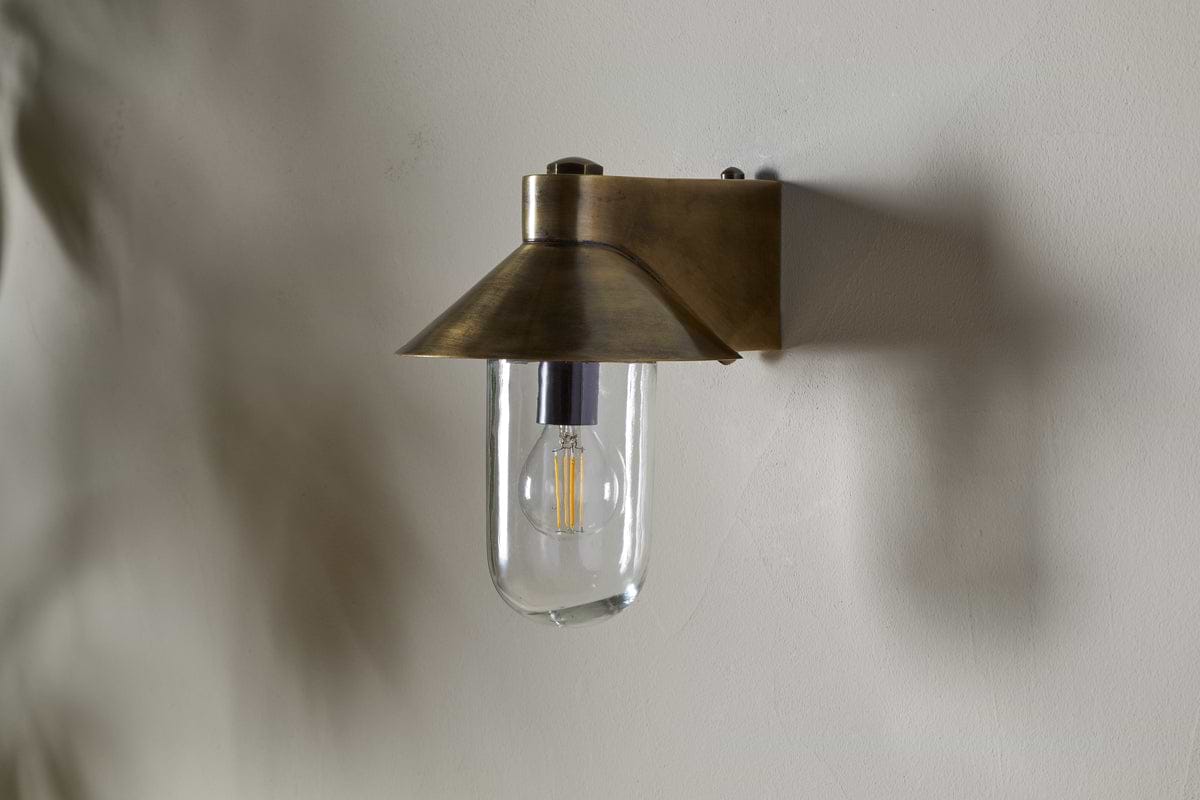 The Singa Outdoor Wall Lamp by nkuku, in antique brass, features a flared conical shade and clear glass that displays an exposed LED filament bulb, casting a soft shadow on the light-colored wall behind it.