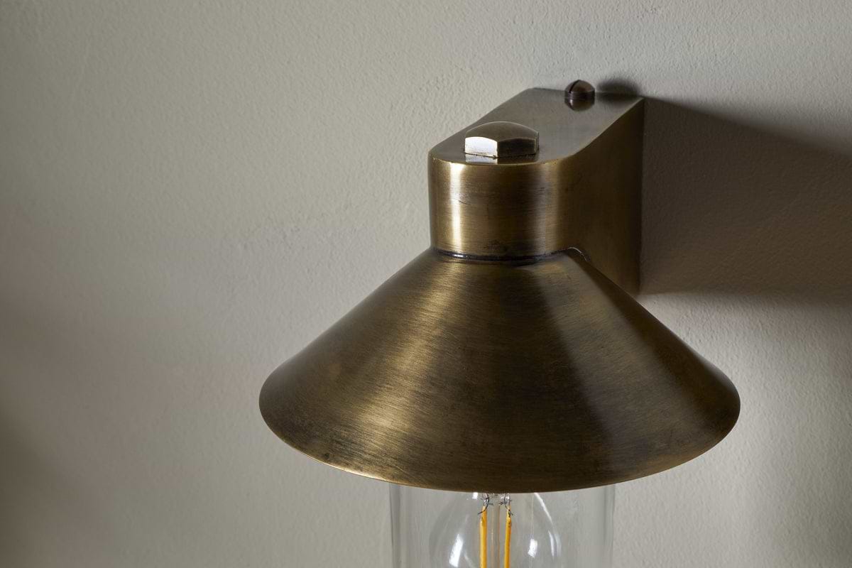 Close-up of nkuku's Singa Outdoor Wall Lamp in antique brass, featuring a wide conical shade and visible bulb. Mounted on a light wall, the fixture casts subtle shadows across the surface.