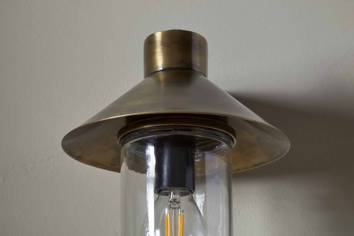 A close-up of the Singa Outdoor Wall Lamp by nkuku in antique brass, featuring a wide conical shade, clear cylindrical glass housing, and visible filament bulb, mounted on a light-colored wall.