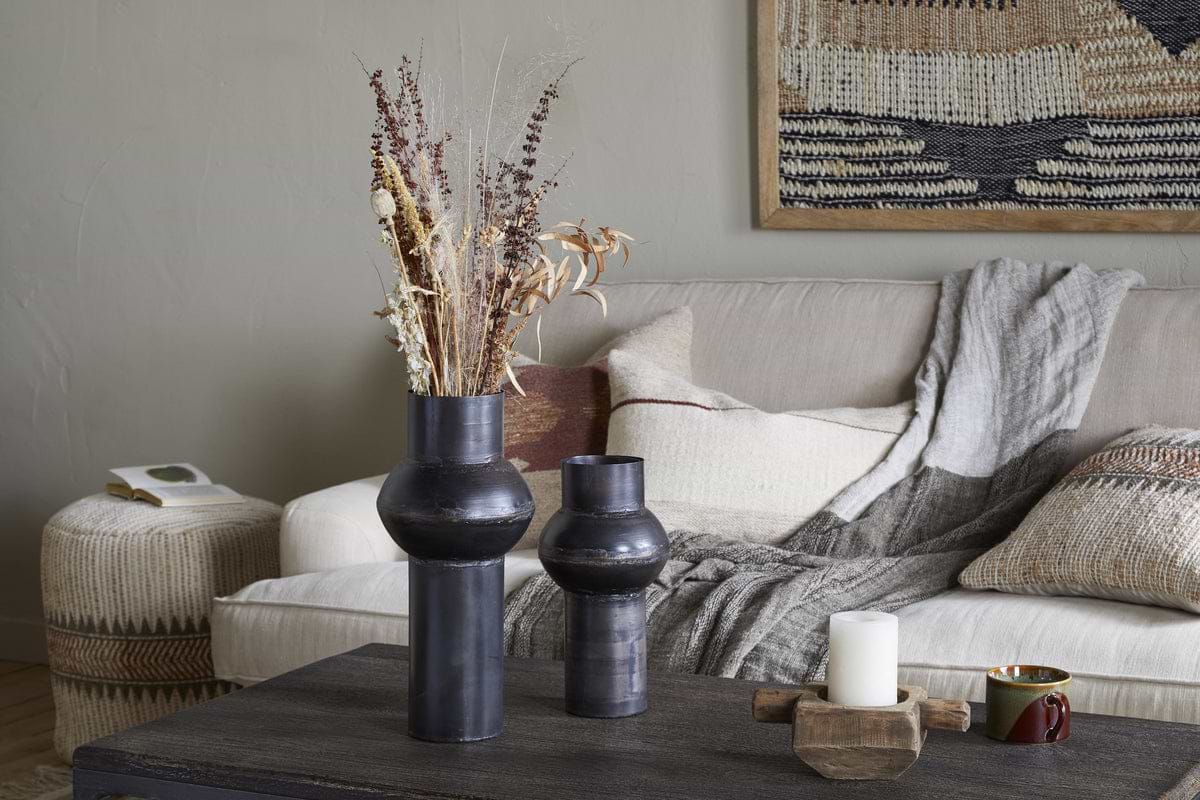A cozy living room with a beige sofa, textured pillows, and a gray throw. On a dark coffee table sits nkuku’s Endo Reclaimed Iron Vase in Aged Black with dried flowers, a white candle on wood, and a cup. A woven basket and tapestry add warmth.