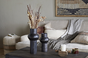 A cozy living room with a beige sofa, textured pillows, and a gray throw. On a dark coffee table sits nkuku’s Endo Reclaimed Iron Vase in Aged Black with dried flowers, a white candle on wood, and a cup. A woven basket and tapestry add warmth.