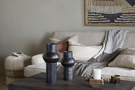 A cozy living room with a beige sofa, gray throw, neutral pillows, woven ottoman with a book, dark coffee table holding an aged bronze vase and the nkuku Endo Reclaimed Iron Vase - Aged Black, two black ceramic vases, a lit white candle, mug, and earth-toned wall tapestry.