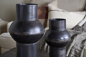Two nkuku Endo Reclaimed Iron Vases in aged black, featuring a dark matte finish, sculptural shape, and visible welded seams, sit on a table. Behind them is a beige sofa with textured and patterned cushions.