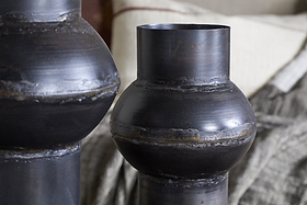 Close-up of two nkuku Endo Reclaimed Iron Vases in aged black, their sculptural forms and visible seams add artisan charm. The rich bronze-black tones complement neutral cushions and a textured gray blanket for a cozy, rustic vibe.