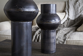 Two Endo Reclaimed Iron Vases in aged black by nkuku, with cylindrical bases and rounded forms, sit on a dark wooden table. A cream sofa with textured pillows and a gray throw blanket creates a cozy living room atmosphere.