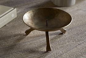 The Kalik Candle Holder by nkuku, crafted in antique brass with three angled legs and a central spike for securing a pillar candle, stands on a textured wooden surface near a beige book and a striped cylindrical object.