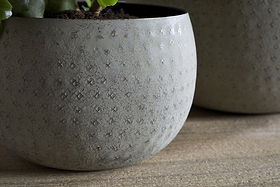 The nkuku Laoko Planter in Olive Grey features a textured, dotted surface and round shape, ideal for modern interiors. A green plant peeks from the top left, with another similar indoor planter softly blurred in the background.