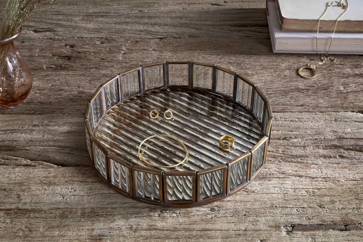 The nkuku Ruaka Glass Vanity Tray in brass sits on a rustic wooden table, displaying gold jewelry—a bracelet, ring, and stud earrings. In the background are stacked books draped with a necklace and a vase of dried flowers.