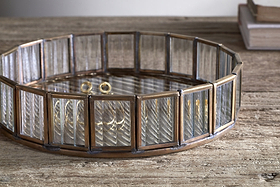 The nkuku Ruaka Glass Vanity Tray – Brass, featuring vertical glass panels in a bronze metal frame, sits on a rustic wooden surface holding gold rings and bracelets, with two stacked books softly lit in the background.
