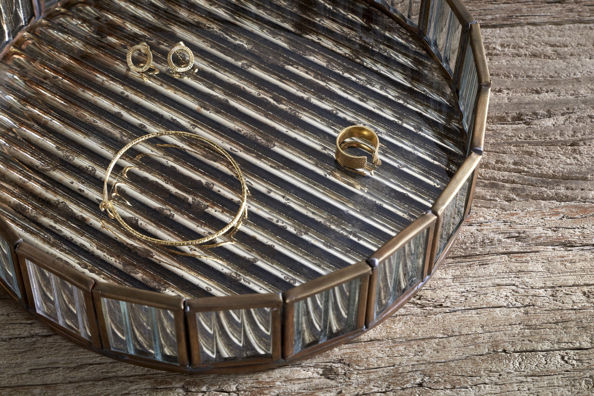 The nkuku Ruaka Glass Vanity Tray - Brass features a round, metallic design with raised edges and a striped, antique mercury glass surface, beautifully displaying gold jewelry on weathered wooden planks.