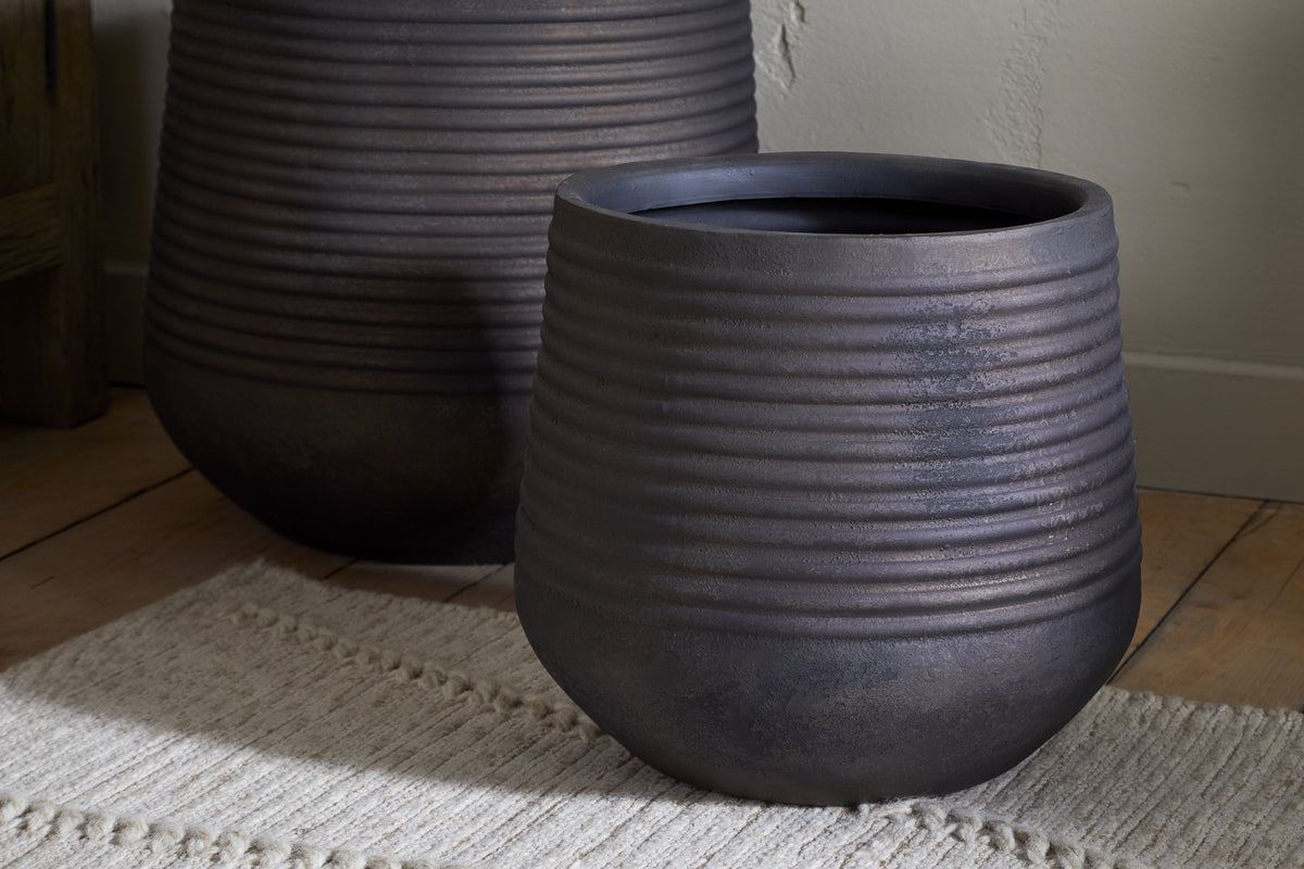 The nkuku Lmaga Metal Grooved Planter in Aged Black stands on a light woven rug atop a wooden floor near a wall, ideal for displaying houseplants. Another similar indoor planter is partially visible in the softly lit background.