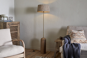 A cozy living room features the nkuku Agua Grooved Floor Lamp - Natural glowing in the corner, with sustainable mango wood furniture, a beige sofa, blue throw, armchair, and textured light walls enhancing the wooden floor.
