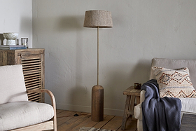 A cozy living room with the nkuku Agua Grooved Floor Lamp in Natural, featuring a textured beige base and matching shade. A sustainable mango wood side table, cream-cushioned armchair, and beige sofa complete the modern space.