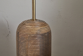 The nkuku Agua Grooved Floor Lamp - Natural features a sustainable mango wood base with horizontal grooves, stacked ring design, and a slender brass rod—ideal for modern spaces against an off-white textured wall.