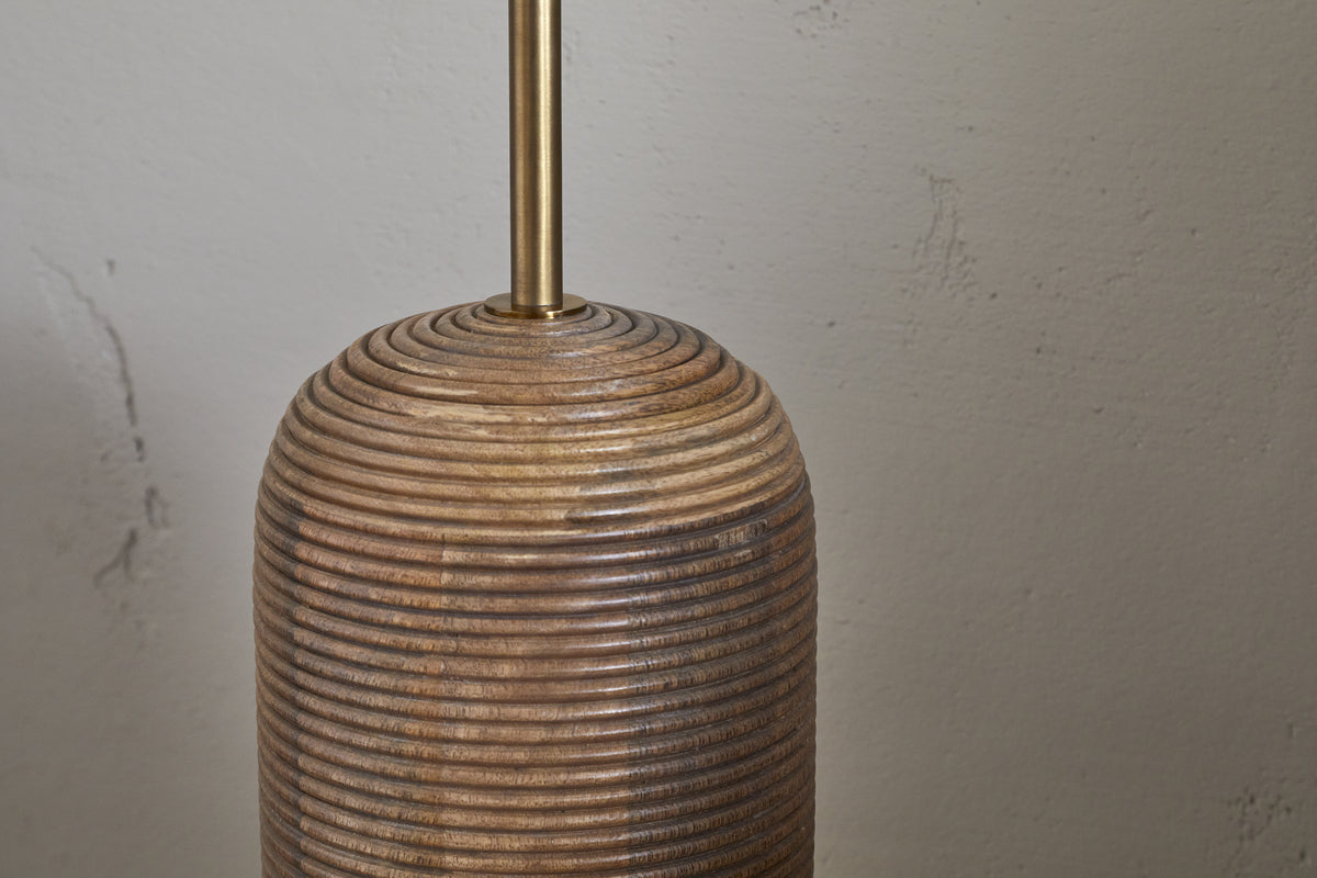 The nkuku Agua Grooved Floor Lamp - Natural features a sustainable mango wood base with horizontal grooves, stacked ring design, and a slender brass rod—ideal for modern spaces against an off-white textured wall.
