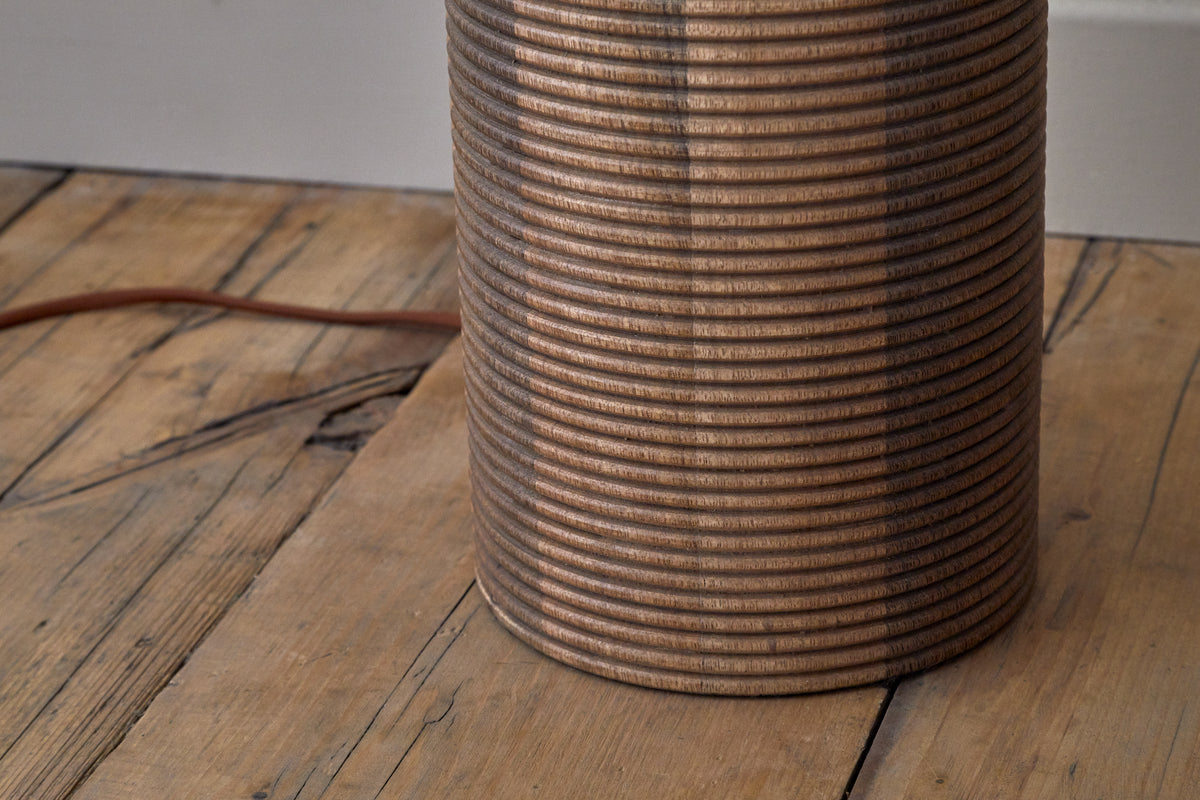 A close-up of the nkuku Agua Grooved Floor Lamp - Natural, crafted from sustainable mango wood, stands on a wooden floor with visible grains. Its brown cord extends behind it—an ideal accent for modern living spaces.