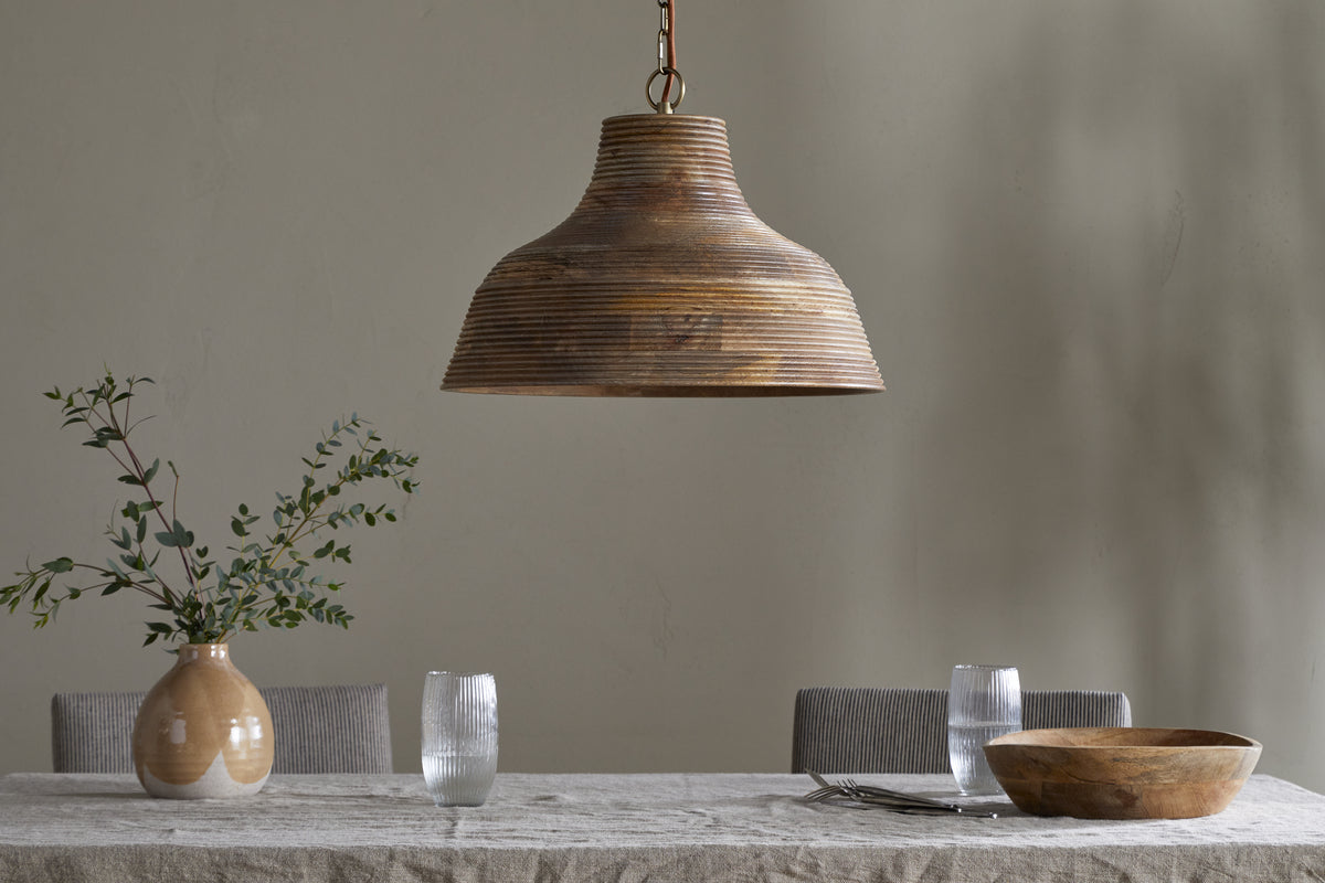 The Nadi Grooved Wood Pendant by nkuku in natural and antique brass hangs above a linen-covered dining table adorned with a beige vase of green branches, ribbed glasses, a wooden bowl, and two forks, set against a light gray wall.
