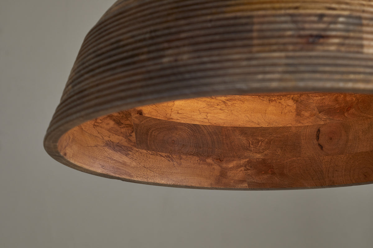 Close-up of nkuku's Nadi Grooved Wood Pendant in Natural & Antique Brass, featuring a ribbed wood exterior and smooth warm-toned interior. A soft glow highlights the wood grain against a neutral background.