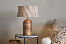 The nkuku Agua Grooved Table Lamp in natural, featuring a textured woven shade, sits on a round gray side table next to a light beige sofa with a patterned cushion. A woven basket is partially visible on the floor behind it.