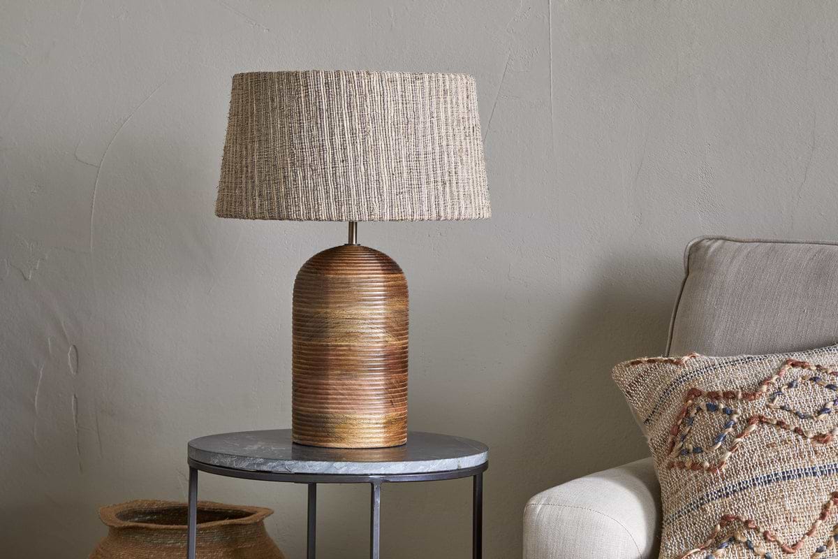 The nkuku Agua Grooved Table Lamp - Natural, featuring a textured woven beige shade, sits on a round dark metal side table with a stone top. Nearby are part of a beige sofa with a patterned pillow and a woven basket by the light gray wall.