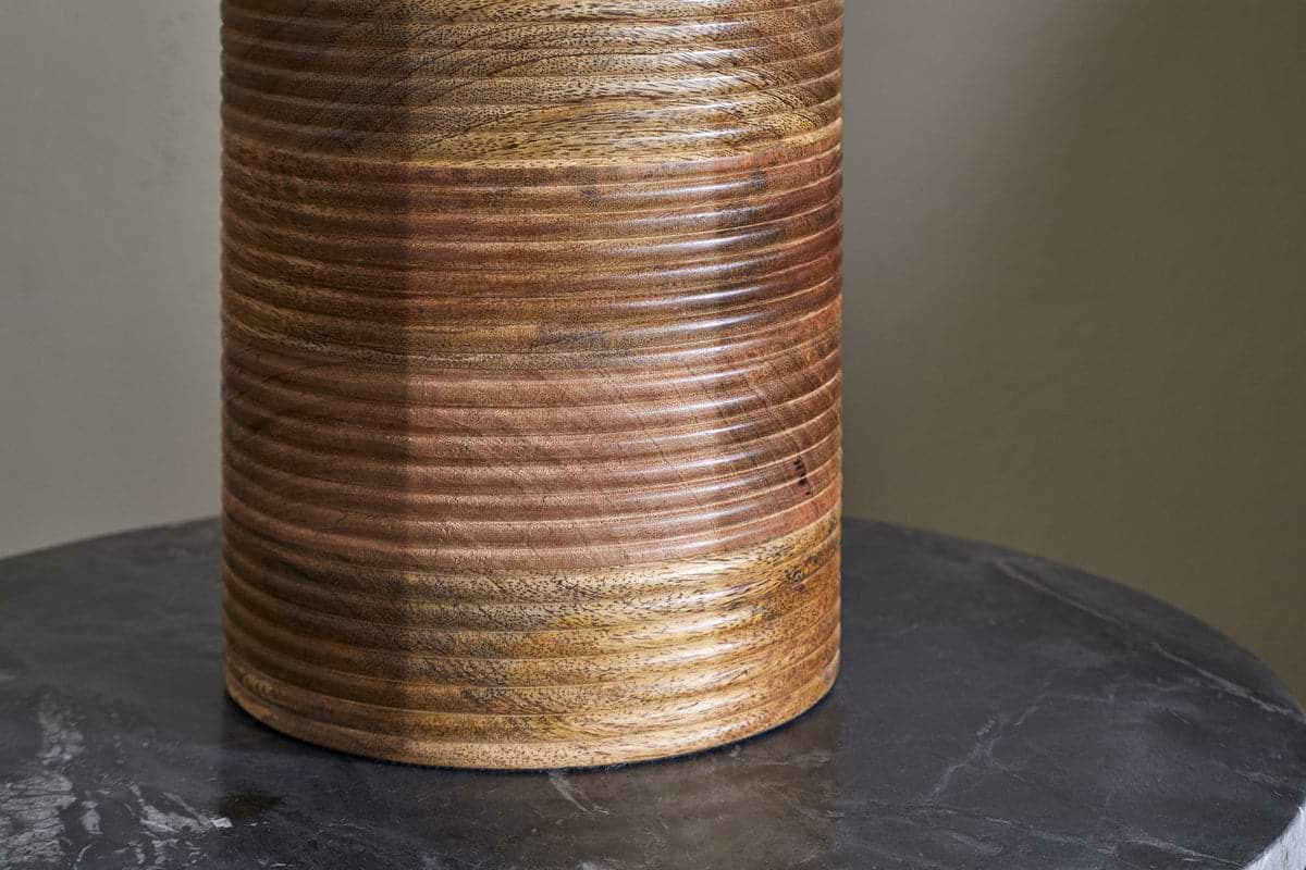 The Agua Grooved Table Lamp - Natural by nkuku features hand-grooved mango wood with horizontal grooves and a smooth, glossy finish. Its unique grain and warm color variations add texture atop a dark, veined marble surface.