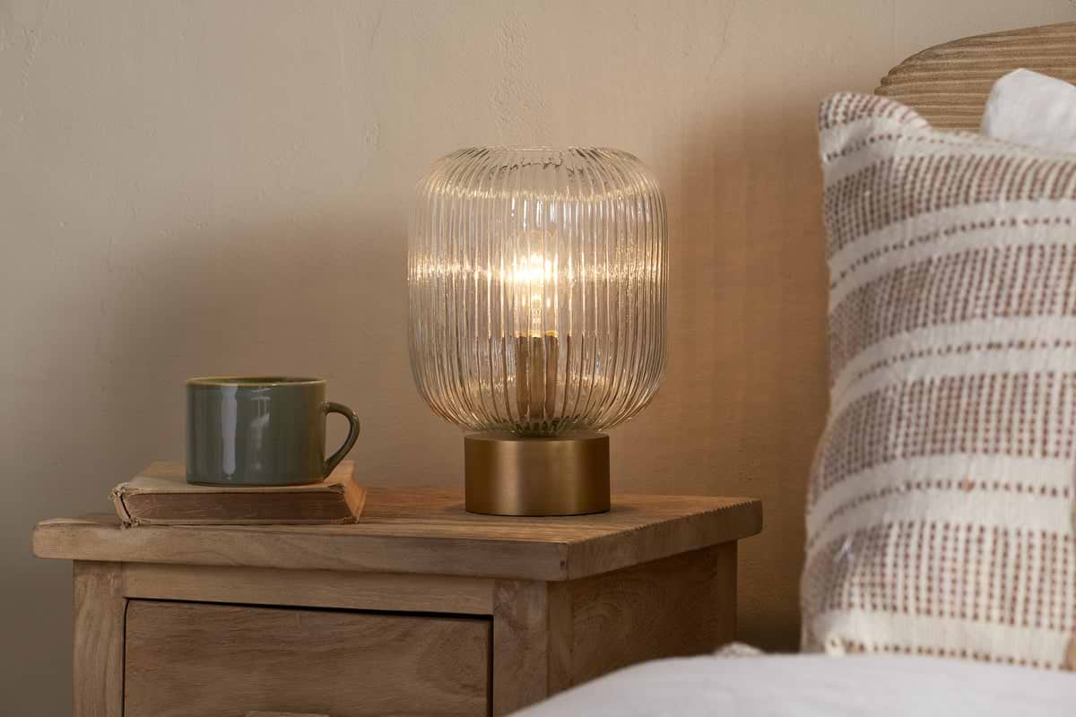The nkuku Rizal Fluted Glass Bedside Table Lamp in Antique Brass & Clear sits on a wooden nightstand next to a green mug on a closed book. A beige and white striped pillow nearby adds to the cozy ambiance with warm lighting.
