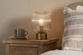 The nkuku Rizal Fluted Glass Bedside Table Lamp in Antique Brass & Clear sits on a wooden nightstand next to a green mug on a closed book. A beige and white striped pillow nearby adds to the cozy ambiance with warm lighting.