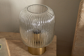 The Rizal Fluted Glass Bedside Table Lamp by nkuku, featuring a cylindrical clear glass design and antique brass base, rests on a wooden surface with sunlight casting soft shadows beside a beige wall and woven chair.