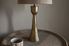 The nkuku Marpha Antique Brass Table Lamp with a beige fabric shade sits on a round wooden tray table, casting accent light. To the left is a white mug with beige and gray stripes, set against a textured off-white wall.