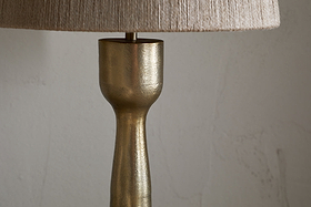 A close-up of the nkuku Marpha Antique Brass Table Lamp shows its textured gold base and beige fabric shade. Ideal as accent lighting, it stands against a plain, light wall with subtle marks. Only the upper base and lower edge of the shade are visible.