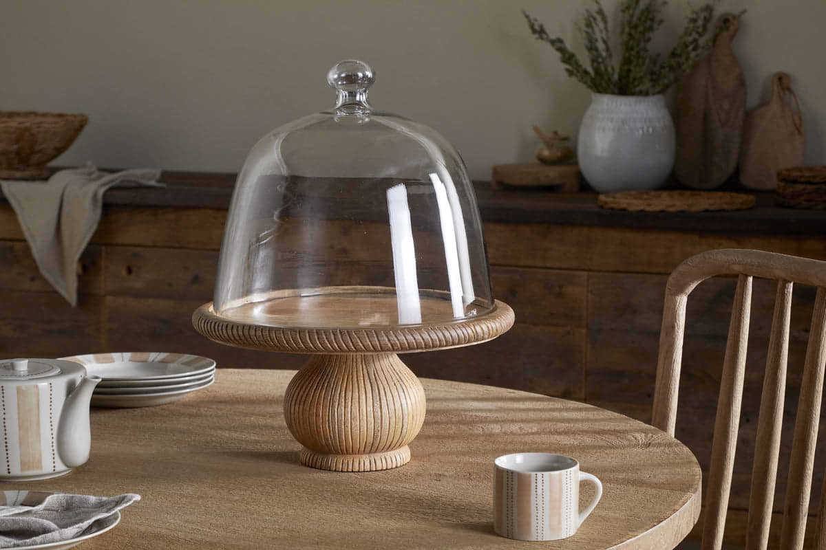 The nkuku Vivan Glass Cake Stand & Dome - Natural sits on a round table, paired with a striped mug, matching plate and teapot, and cloth napkin. Rustic decor and ceramics accent the wooden sideboard behind.
