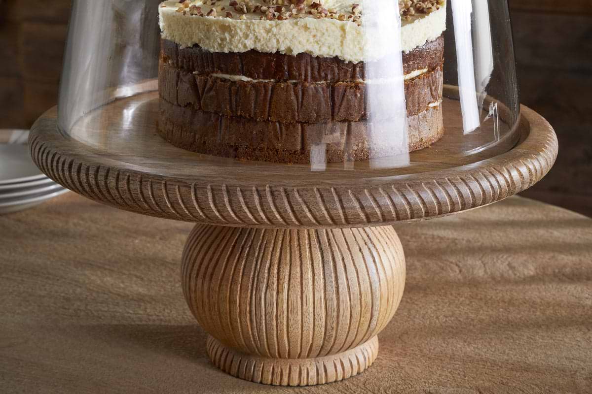 A layered cake with white frosting and nuts sits on the nkuku Vivan Glass Cake Stand & Dome - Natural, featuring a carved mango wood base and clear glass cover, with serveware stacked in the blurred background.