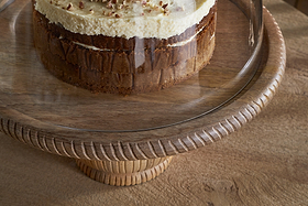 A partially visible layered cake with cream frosting and nut crumbles sits on the nkuku Vivan Glass Cake Stand & Dome - Natural, featuring a grooved mango wood base and glass cover, placed on a wooden table.