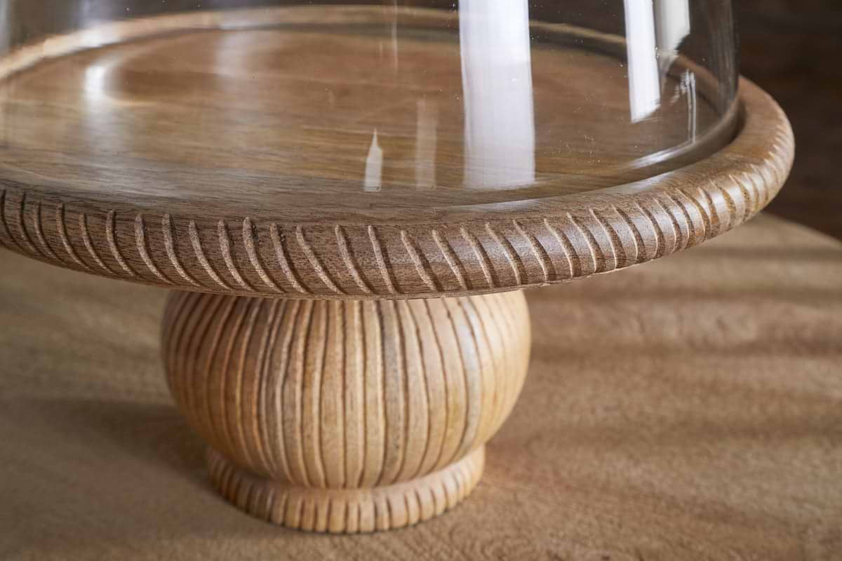 The Vivan Glass Cake Stand & Dome – Natural by nkuku features a mango wood base with carved texture and fine grain, topped with a clear dome, showcasing the light finish against a softly blurred background.