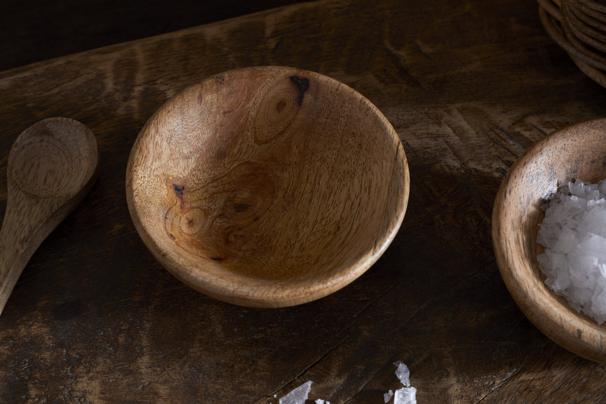 A close-up of the nkuku Daruka Salt & Pepper Dipping Bowl with Spoon in natural wood rests on a rustic surface, flanked by additional wooden spoons and a bowl of coarse sea salt—perfect for charming, artisanal table settings.
