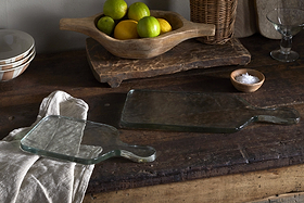 Two nkuku Komal Serving Platters in clear glass with handles rest on a rustic wooden table. One sits atop a beige cloth. Nearby are bowls, a wooden fruit bowl with lemons and limes, a wicker basket, and serveware.