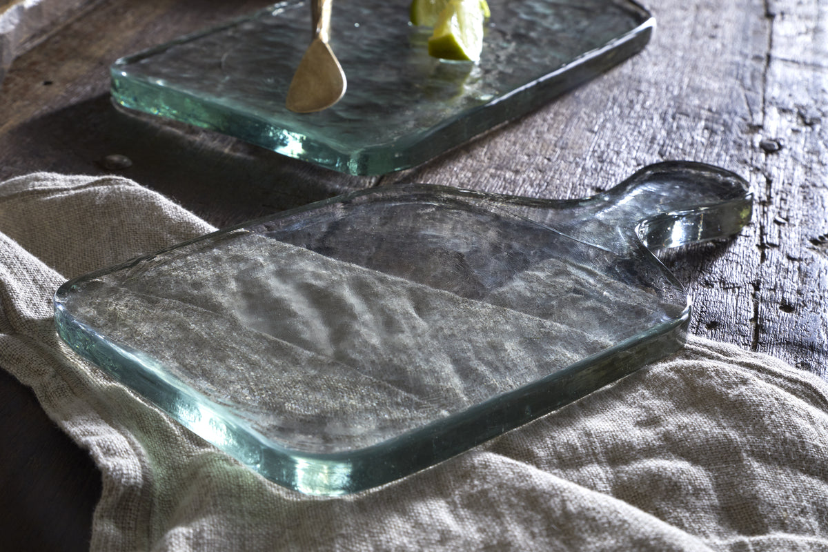 The Komal Serving Platter - Clear by nkuku, a thick glass board with handle, sits on a beige cloth atop a rustic wooden table. In the background are another platter, a wooden spoon, and a lime wedge, all softly lit by natural light.