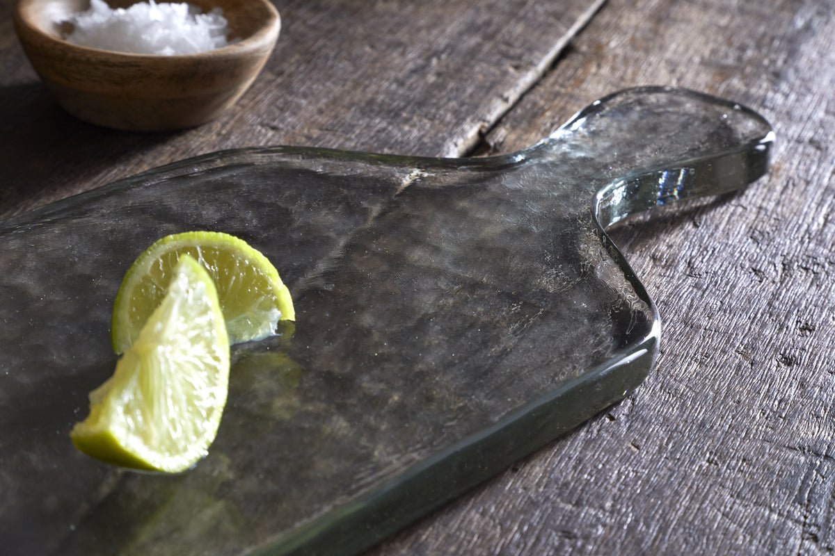 Two lime wedges rest on the nkuku Komal Serving Platter - Clear, placed on a rustic wooden surface. A small wooden bowl with coarse sea salt adds contrast, while natural light accentuates the platter and fresh limes.