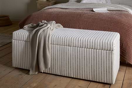 The nkuku Sanja Stripe Cotton & Linen Upholstered Storage Bench in Grey sits at the foot of a bed with a textured reddish-brown quilt and white sheets, adding practical bedroom storage to the cozy, minimalist space with neutral decor and wooden floors.