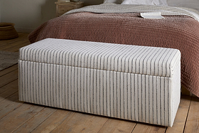 The nkuku Sanja Stripe Cotton & Linen Upholstered Storage Bench - Grey, featuring vertical beige and grey stripes, adds stylish storage at the foot of the bed with mauve-brown quilt and white bedding in a cozy bedroom.