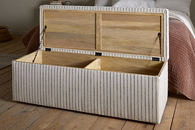 The nkuku Sanja Stripe Cotton & Linen Upholstered Storage Bench in grey features a hinged lid, two wooden compartments, and light fabric with thin gray stripes, offering stylish bedroom storage beside a bed.