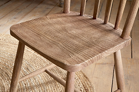 A close-up of the nkuku Om Mango Wood Dining Chair in natural finish shows its textured seat and straight back spindles. The chair stands on a light wood floor next to a round woven jute rug, creating a rustic, airy atmosphere.