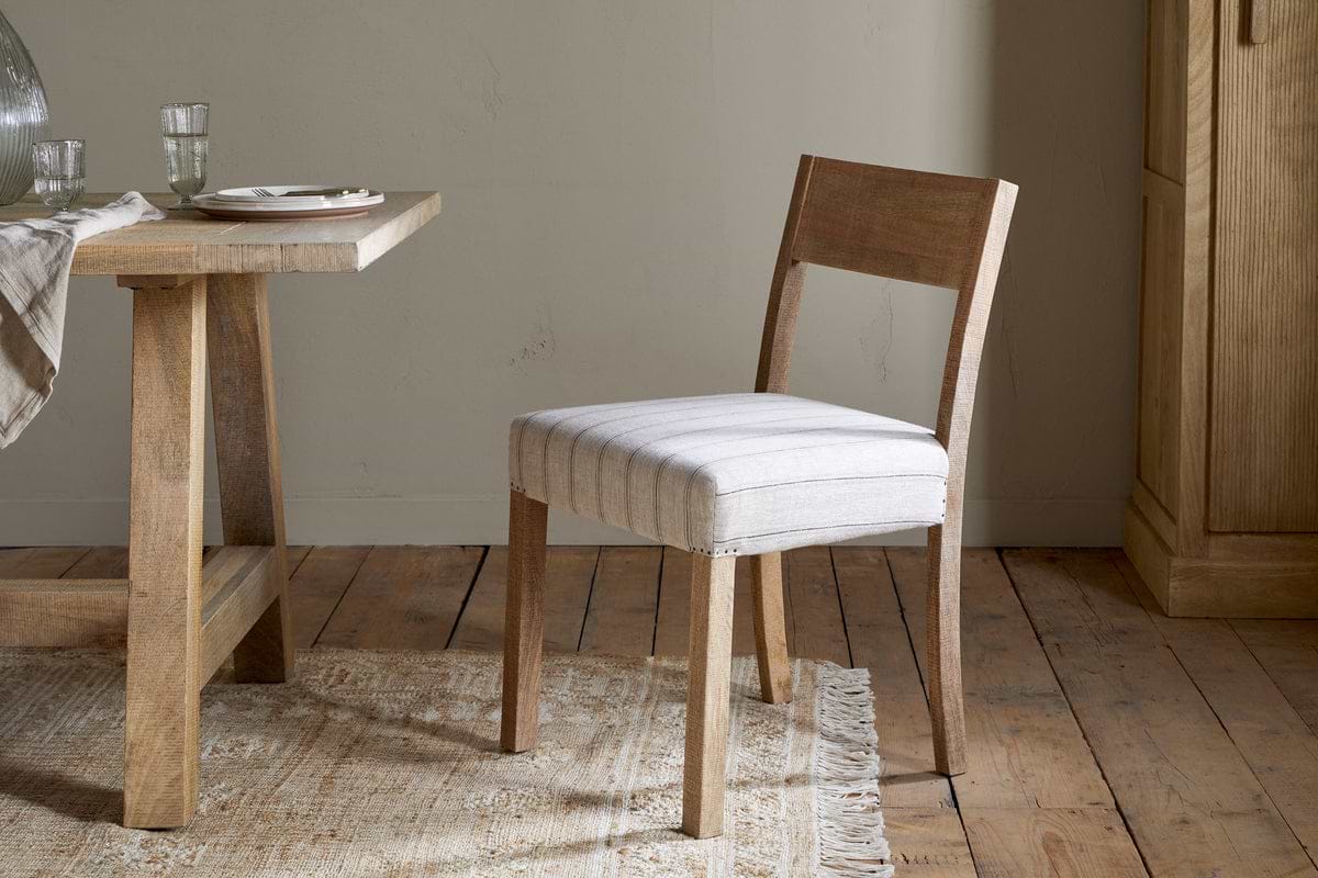 The nkuku Nidhi Linen Upholstered Dining Chair in natural mango wood with off-white striped linen stands by a matching table set on a woven rug atop a rustic wooden floor, with plates, glasses, and a napkin arranged neatly on the tabletop.