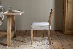 The nkuku Nidhi Linen Upholstered Dining Chair in natural mango wood sits beside a matching table set with plates, glasses, and a striped runner, all arranged on a fringed beige rug over wooden floorboards by a light wall.