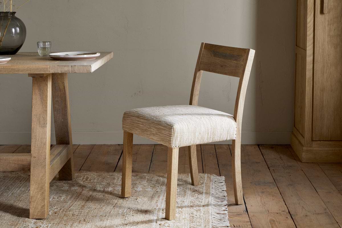 The Zoha Cotton & Jute Upholstered Dining Chair - Natural by nkuku sits beside a matching table set with plates and a glass, on a fringed rug over rustic wood flooring, with a cabinet and neutral wall in the background.